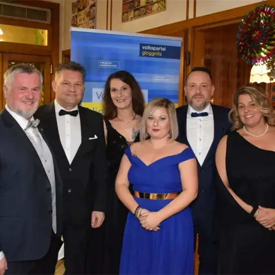 Eine Gruppe von sechs Personen, drei Männer und drei Frauen, in formeller Kleidung, posiert für ein Foto vor einem dekorierten Hintergrund.