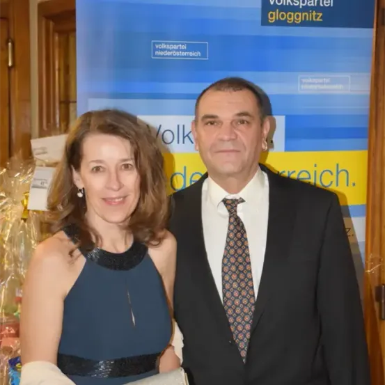 Ein Mann und eine Frau stehen nebeneinander vor einem blauen Banner, auf dem 'volkspartei gloggnitz' steht. Der Mann trägt einen Anzug und eine Krawatte, während die Frau ein blaues Kleid trägt. Sie lächeln beide.