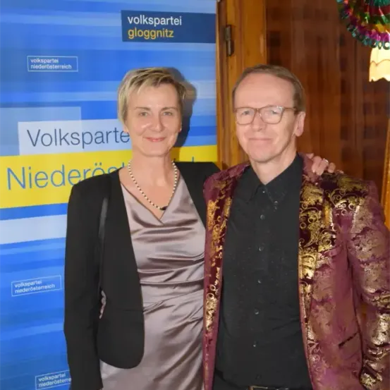 Zwei Personen stehen nebeneinander vor einem Banner mit der Aufschrift 'Volkspartei Niederosterreich'. Die Frau trägt eine Halskette und eine schwarze Jacke, der Mann trägt eine Brille und ein aufwendiges Hemd.