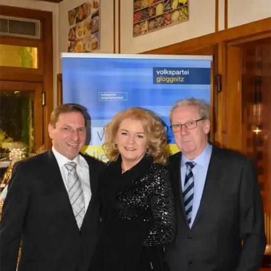 Drei formell gekleidete Personen stehen eng beieinander vor einem Banner und lächeln für ein Foto. Das Banner hat blauen und weißen Text.