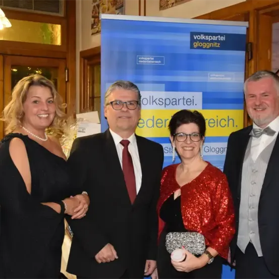 Vier Menschen in formeller Kleidung stehen vor einem Banner mit dem Text 'Volkspartei gloggnritz'. Die Frau links trägt ein schwarzes Kleid, während die andere Frau eine Handtasche hält. Die beiden Männer tragen Anzüge und Krawatten.