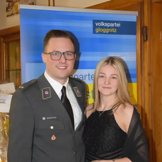Ein Mann in Uniform steht neben einer Frau in einem schwarzen Kleid. Sie lächeln beide und stehen vor einem Schild mit den Worten 'Volkspartei gloggnitz'.