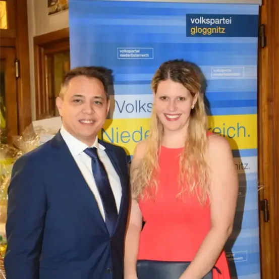 Ein Mann und eine Frau stehen neben einem blau-gelben Banner mit Text in Weiß und Orange. Sie lächeln beide.