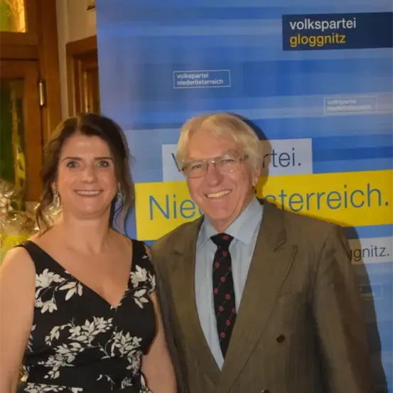 Zwei Personen, eine Frau und ein Mann, stehen dicht nebeneinander vor einem Banner. Die Frau trägt ein schwarzes Blumenkleid, und der Mann ein braunes Kostüm mit Krawatte. Sie lächeln beide in die Kamera.