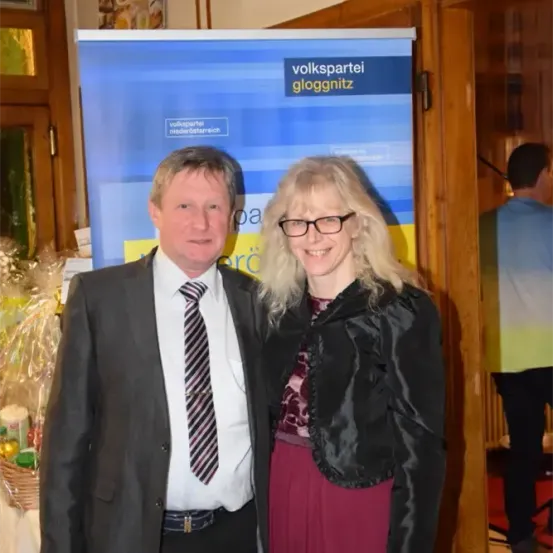 Ein Mann und eine Frau stehen neben einem blauen Banner mit dem Text 'volkspartei gloggnitz'. Der Mann trägt einen Anzug und eine Krawatte, und die Frau trägt eine Brille und eine schwarze Jacke. Hinter ihnen ist eine teilweise sichtbare Person. Ein Korb mit Gegenständen und eine Holztür sind ebenfalls zu sehen.