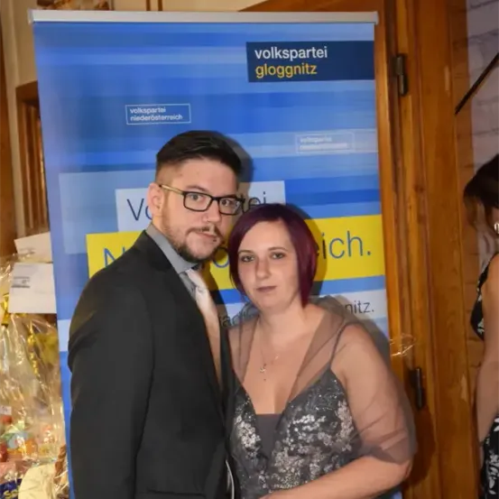 Ein Mann und eine Frau stehen eng nebeneinander vor einem Volkspartei Gloggnitz Banner. Der Mann trägt einen Anzug und eine Brille, während die Frau rote Haare und ein silbernes Kleid hat.