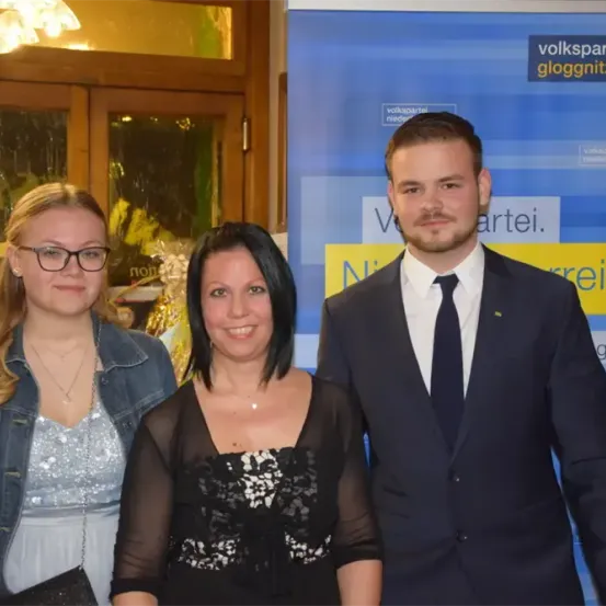 Drei Personen stehen zusammen vor einem blauen Banner mit gelbem Text. Ein Mann und zwei Frauen lächeln für ein Foto.