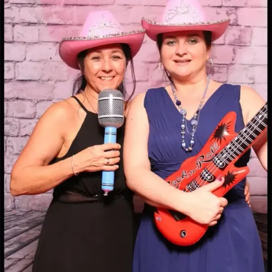 Zwei Frauen in rosa Cowboyhüten stehen nebeneinander, eine hält ein Mikrofon und die andere eine Gitarre. Sie lächeln beide in die Kamera.