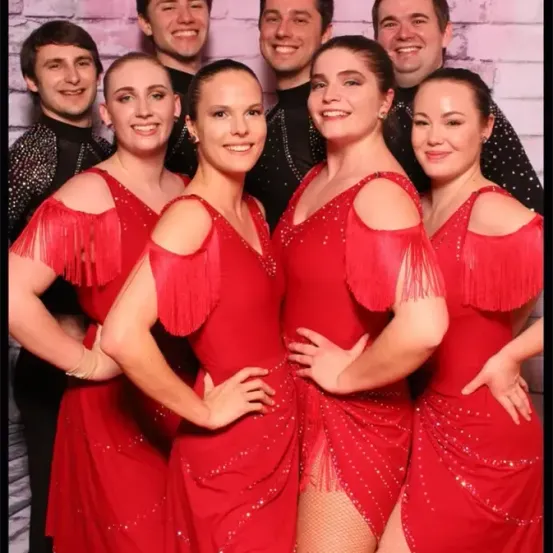 Eine Gruppe von sechs Personen, vier Frauen und zwei Männer, in roten Kleidern mit Fransen und Pailletten, lächeln und posieren für ein Foto.