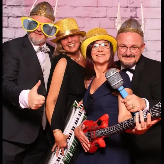 Vier Erwachsene posieren für ein Foto, tragen verschiedene Kostüme und halten Requisiten wie eine Gitarre und ein Keyboard. Der Mann auf der linken Seite gibt einen Daumen hoch.