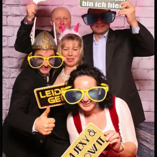 Fünf Personen posieren für ein Foto mit verschiedenen Requisiten. Eine Frau hält ein Schild mit der Aufschrift 'I'm Jexy Now It.' Eine andere Frau hat ein Schild mit der Aufschrift 'Leiden.' Ein Mann im Anzug hält ein Schild mit der Aufschrift 'Wegen dem Bier bin ich hier.' Ein Mann in einem Helm und einer in Hasenohren sind ebenfalls anwesend.