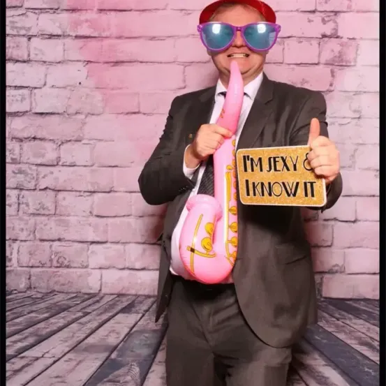 Ein Mann in Anzug hält eine rosa aufblasbare Saxophon, trägt einen roten Hut und eine Sonnenbrille, vor einer Backsteinmauer. Er gibt einen Daumen hoch.