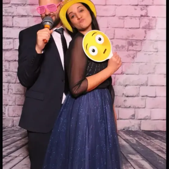 Ein Mann und eine Frau posieren für ein Foto mit einem Ausschnitt eines lächelnden Gesichts, der Mann hält ein Mikrofon und die Frau trägt einen gelben Hut. Sie stehen vor einer rosa Ziegelmauer.