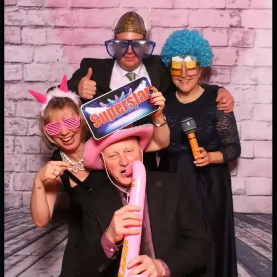 Vier Menschen posieren für ein Foto. Sie tragen Hüte, Brillen und ein Mikrofon. Der Mann in der Mitte bläst in einen rosafarbenen Ballon. Hinter ihnen ist eine Ziegelmauer.