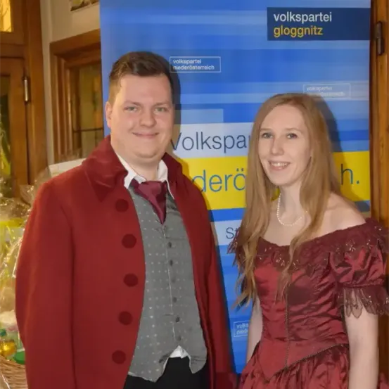 Ein Mann und eine Frau in roten Kleidern, die lächeln, stehen vor einem blauen Banner mit Text und einem Korb mit Gegenständen dahinter.