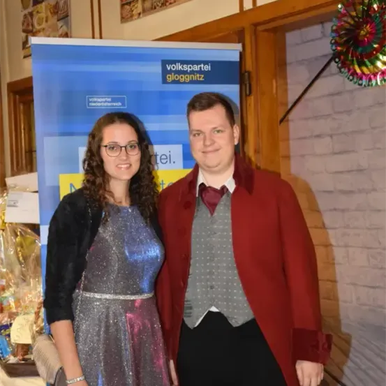 Ein Mann und eine Frau stehen in einem festlichen Ambiente eng beieinander. Die Frau trägt ein glänzendes Kleid und der Mann einen roten Mantel. Sie stehen vor einem Schild mit 'volkspartei gloggnitz' darauf. Hinter ihnen befindet sich eine Ziegelwand und eine bunte Dekoration.