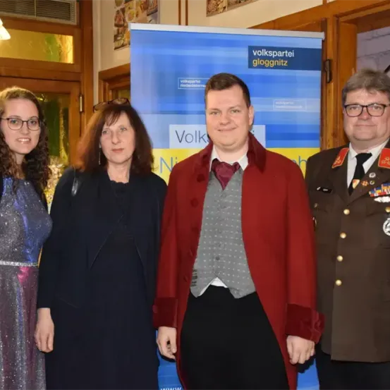 Vier Personen stehen nebeneinander, wahrscheinlich für ein Foto. Der Mann in der Mitte trägt einen roten Mantel. Der Mann auf der rechten Seite ist in einer Militäruniform. Ein blauer Banner mit weißem Text ist hinter ihnen.