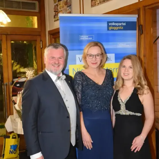 Drei Personen stehen vor einem blauen Banner mit der Aufschrift 'volkspartei gloggnitz'. Der Mann trägt einen schwarzen Anzug und ein weißes Hemd, während die beiden Frauen Abendkleider tragen. Hinter ihnen befindet sich ein gelber Beutel mit der Aufschrift 'Ich liebe NO'.