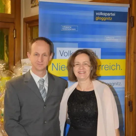 Ein Mann und eine Frau stehen nebeneinander vor einem blauen Banner, auf dem 'Volkspartei gloggnitz Niederoesterreich' steht.