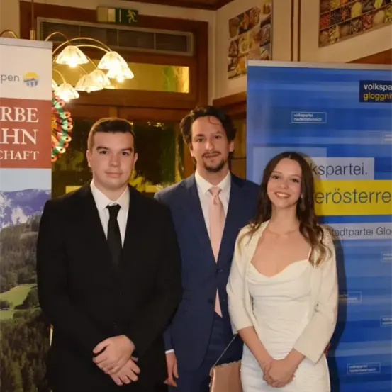 Drei Menschen in formeller Kleidung stehen nebeneinander vor einer Kulisse mit Berglandschaft und einem gelben Banner mit deutschem Text.