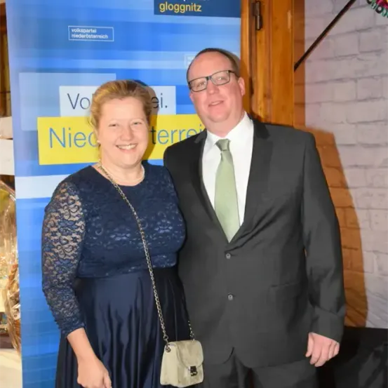 Ein Mann und eine Frau stehen in formeller Kleidung nebeneinander. Die Frau trägt ein blaues Kleid mit Spitzendetails und der Mann ist in einem schwarzen Anzug mit einer grünen Krawatte. Sie lächeln und stehen vor einem blauen und gelben Hintergrund.