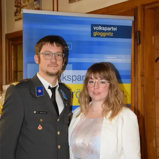 Ein Mann und eine Frau stehen vor einem Banner. Der Mann ist in Uniform und trägt eine Brille. Die Frau lächelt und trägt ebenfalls eine Brille.