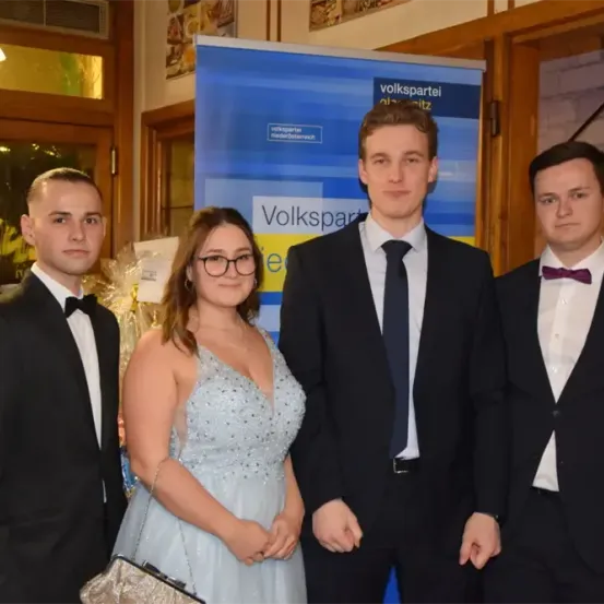 Vier Menschen in formeller Kleidung posieren für ein Foto vor einem blauen Banner mit Text. Die Frau trägt ein funkelndes Kleid, und die Männer tragen Krawatten und Fliegen.