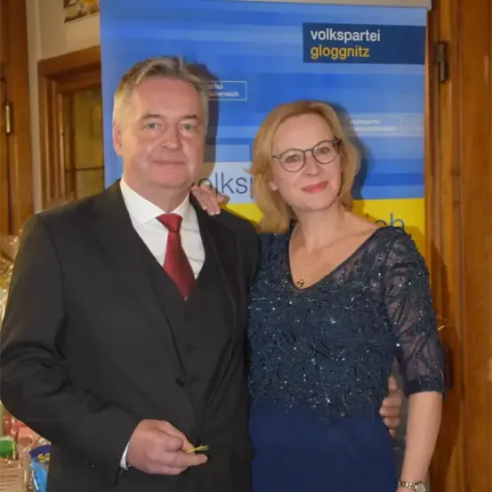 Ein Mann und eine Frau stehen vor einem blauen Banner mit dem Text 'volkspartei gloggnitz'. Der Mann trägt einen Anzug und eine rote Krawatte, und die Frau trägt ein blaues Kleid und eine Brille.