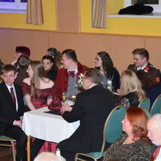 Eine Zusammenkunft von Menschen in formeller Kleidung in einem Raum, einige sitzend und andere stehend, wobei zwei Männer in Uniformen in der Nähe eines Tisches mit Blumen und Getränken sprechen.