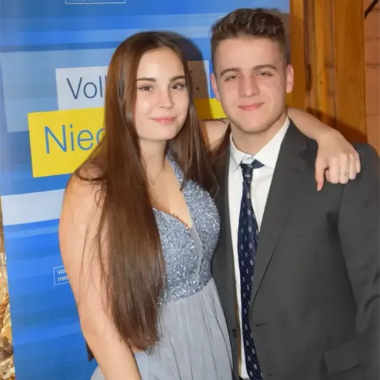 Ein junges Paar lächelt für ein Foto vor einem blauen Hintergrund mit Text. Der Mann trägt einen Anzug und eine Krawatte, während die Frau ein silbernes Kleid trägt.