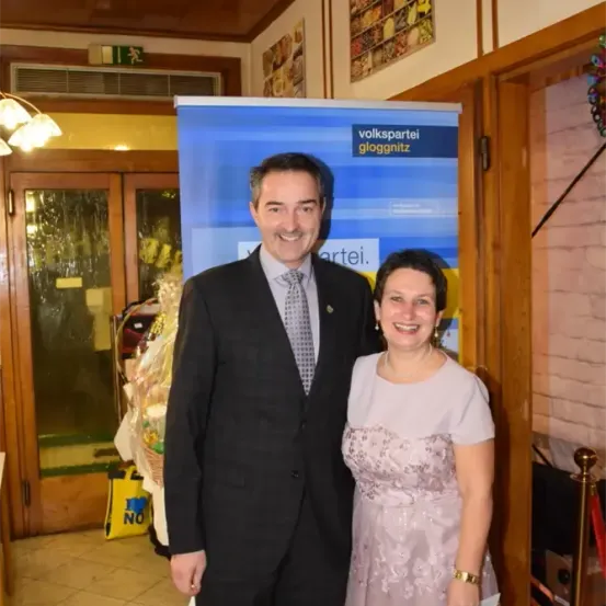 Ein Mann und eine Frau stehen lächelnd vor einem blauen Banner mit der Aufschrift Volkspartei Gloggnitzer. Dahinter führt eine Tür zu einem Raum mit einem Korb und einer gelben Tasche.