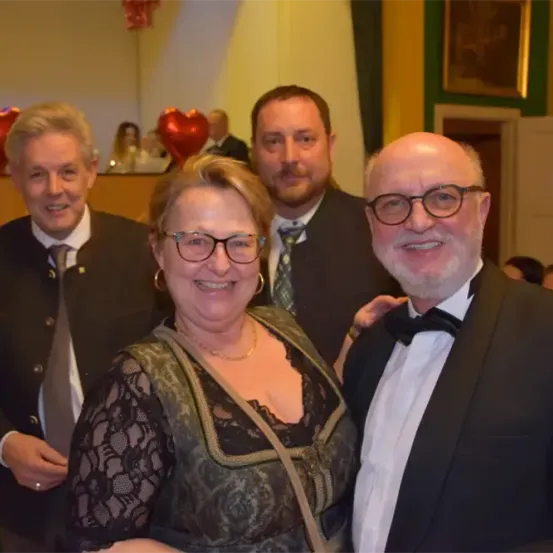 Vier Personen, zwei Männer und zwei Frauen, posieren lächelnd für ein Foto. Sie sind formell gekleidet. Hinter ihnen befinden sich rote Herzdekorationen und ein Bilderrahmen an der Wand.