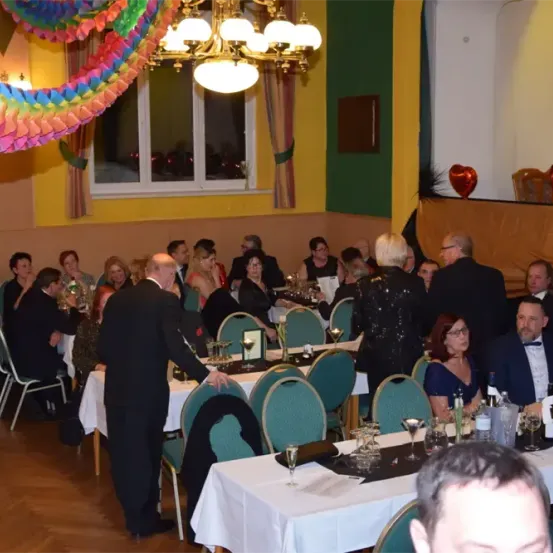 Eine Gruppe von Menschen in formeller Kleidung versammelt sich zu einer Feier in einem Bankettsaal. Es gibt Tische mit Gläsern, Flaschen und Weingläsern. Der Raum hat eine Kronleuchter, ein Fenster und eine bunte Dekoration an der Decke.