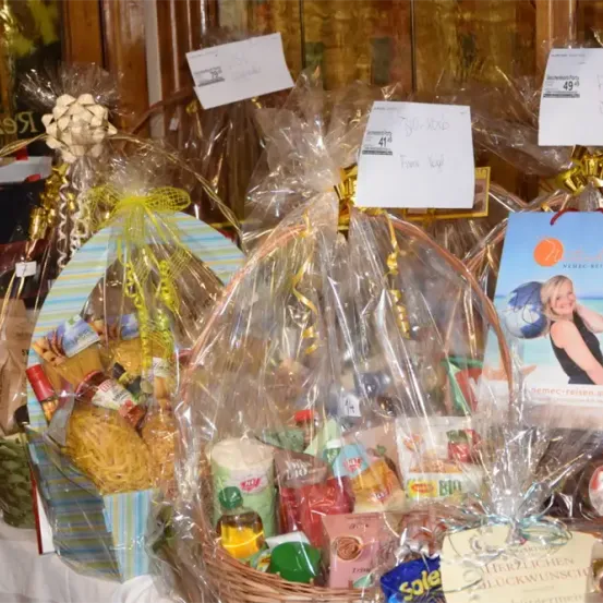 Mehrere Geschenkkörbe mit verschiedenen Lebensmitteln, in Plastik verpackt und mit Bändern gebunden. Einige haben Preisschilder. Ein Poster mit einer Frau und Text ist sichtbar.