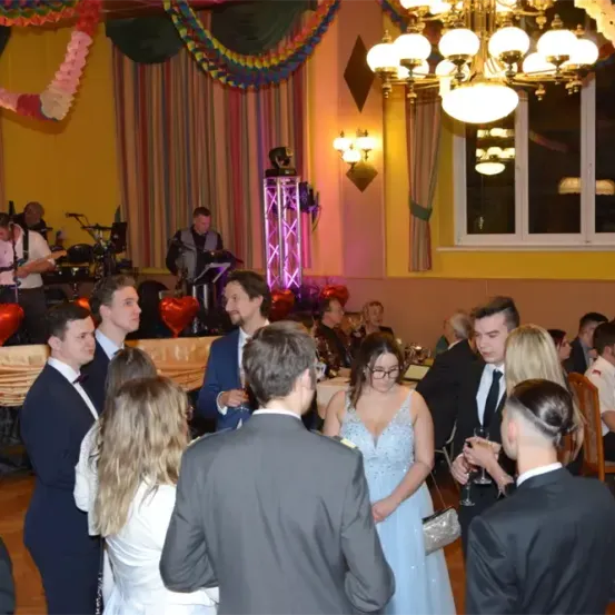 Eine Gruppe von Menschen versammelt sich in einem Bankettsaal, einige unterhalten sich und andere halten Gläser. Der Saal ist mit Luftballons dekoriert und eine Bühne ist mit einer Band eingerichtet, die spielt.