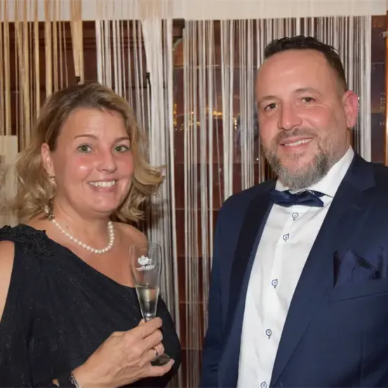 Ein Mann und eine Frau in formeller Kleidung lächeln in die Kamera. Die Frau hält ein Glas Champagner.