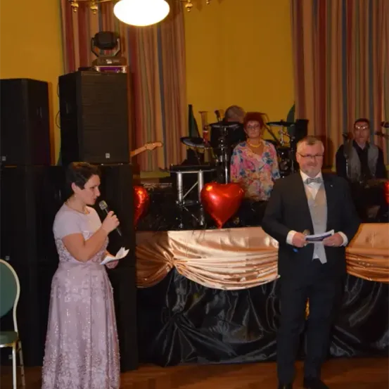 Eine Frau in einem Kleid hält ein Mikrofon in der Hand und steht vor einer Bühne mit einem großen roten Herz und einer Band. Ein Mann in einem Anzug steht rechts von ihr und hält ebenfalls ein Mikrofon und Papier.