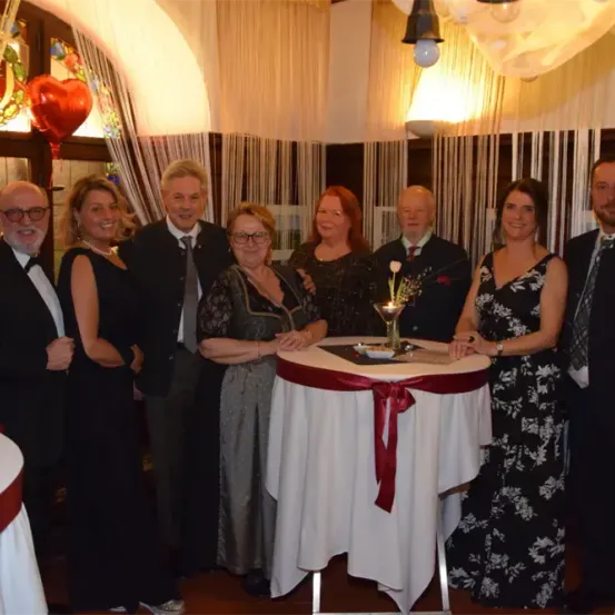 Eine Gruppe von Menschen, einschließlich Erwachsener in formeller Kleidung, steht um einen Tisch mit Blumenarrangement und lächelt für ein Foto.