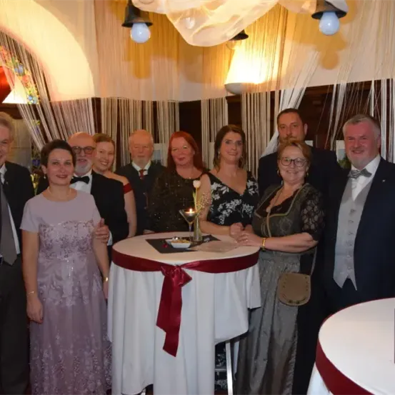 Eine Gruppe von Menschen in formeller Kleidung versammelt sich um einen Tisch in einem Raum mit dekorativen Lichtern. Eine Frau in einem Kleid steht in der Mitte, mit einem roten Band und einer Kerze auf dem Tisch. Dahinter hängt ein herzförmiger Ballon an der Decke.