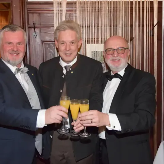 Drei Männer in formeller Kleidung lächeln und posieren für ein Foto, während sie Sektgläser halten. Der Mann auf der linken Seite trägt eine Fliege und eine Abzeichen. Der Mann in der Mitte trägt eine Krawatte. Der Mann auf der rechten Seite trägt eine Fliege und einen Ring. Sie stehen vor einem weißen Vorhang.