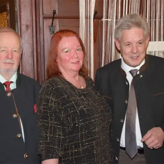 Drei Personen stehen eng beieinander in einem formellen Umfeld. Die Frau hat rotes Haar, und die beiden Männer tragen Anzüge und Krawatten. Sie alle lächeln.