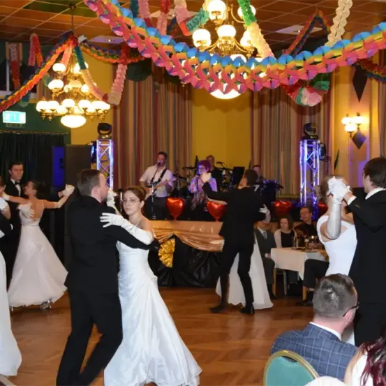 Eine lebhafte Hochzeitsfeier mit einer Band, die hinter einem dekorierten Tisch spielt. Eine Braut und ein Bräutigam tanzen in der Mitte, während andere Gäste um sie herum tanzen. Der Raum ist mit bunten Bändern und Lichtern dekoriert.