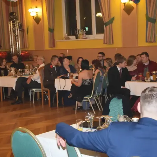 Eine Gruppe von Menschen sitzt an Tischen in einem Bankettsaal. Einige trinken aus Gläsern. Ein Mann in einem blauen Anzug steht, möglicherweise spricht er die Gruppe an. Der Saal hat Holzböden, gelbe Wände und eine Bühne mit einem Lautsprecher.