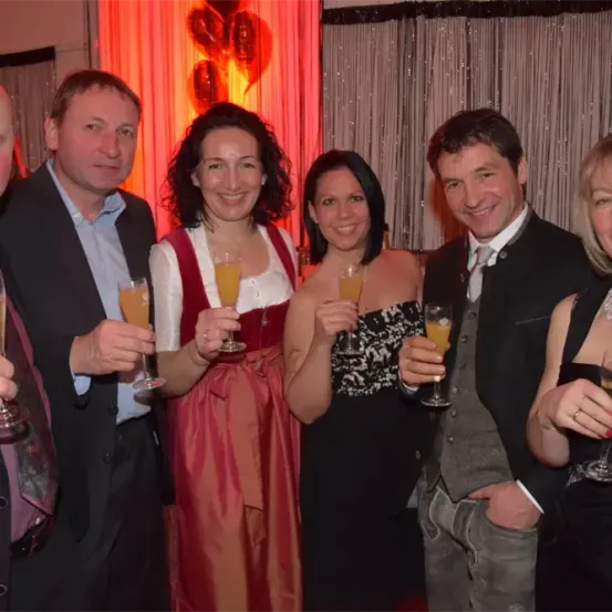 Sechs Personen, drei Männer und drei Frauen, stehen zusammen und halten Weingläser. Sie alle lächeln und scheinen sich auf einer Feier zu befinden. Der Hintergrund besteht aus roten Vorhängen und einem dekorativen Hintergrund.