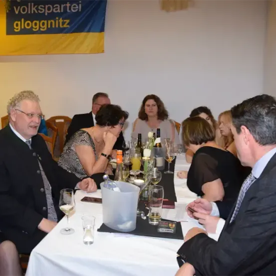 Eine Gruppe von Menschen sitzt um einen Tisch herum, mit Weingläsern und Flaschen vor ihnen. Sie befinden sich wahrscheinlich bei einer formellen Veranstaltung, wobei ein Mann eine Brille und einen Anzug trägt.