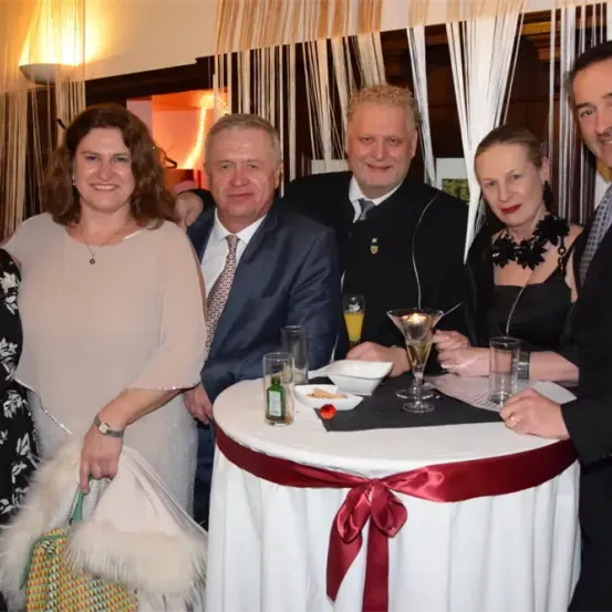 Eine Gruppe von Menschen, darunter Männer und Frauen, steht bei einer Veranstaltung um einen Tisch herum. Sie sind formell gekleidet und lächeln. Auf dem Tisch befinden sich Gläser, eine Schüssel und eine Flasche. Hinter ihnen sind ein Vorhang und eine Lampe zu sehen.