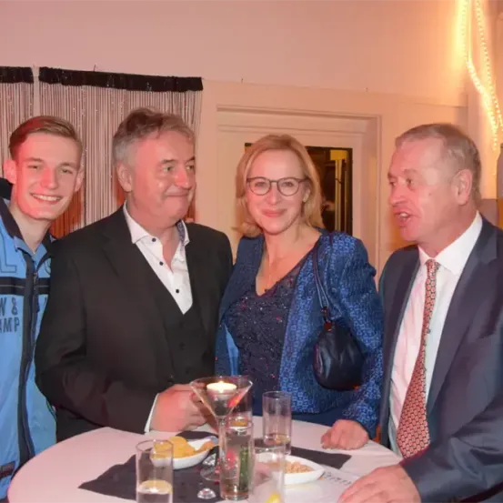 Vier Personen sitzen lächelnd und unterhaltend um einen Tisch herum. Der Mann auf der linken Seite trägt eine blaue Jacke mit Text, während die anderen formell gekleidet sind. Auf dem Tisch stehen eine brennende Kerze, eine Schüssel mit Essen und Gläser. Ein Tür und ein Vorhang sind im Hintergrund zu sehen.