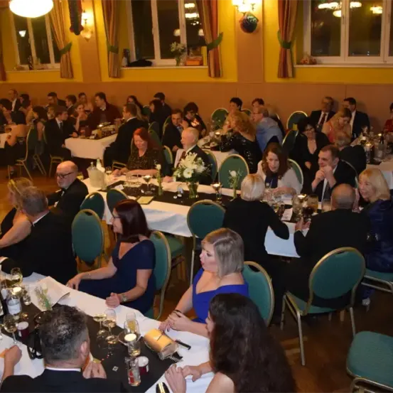 Eine große Gruppe von Menschen sitzt in einem Bankettsaal um Tische herum. Sie sind in formeller Kleidung gekleidet. Auf den Tischen stehen Kerzen, Blumen und Weingläser.