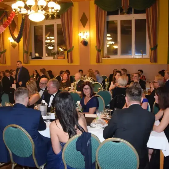 Eine formelle Veranstaltung mit Menschen an Tischen, einige stehen und unterhalten sich. Flaschen, Weingläser und Teller sind auf den Tischen. Der Raum ist mit bunten Girlanden und Vorhängen dekoriert.