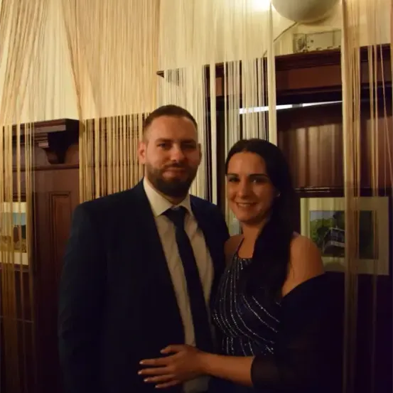 Ein Mann und eine Frau posieren für ein Foto vor einer Holzwand mit Bilderrahmen und einer Lichtarmature.